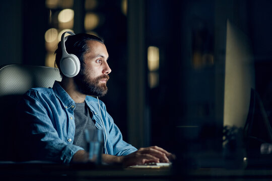 Focused hipster young man with beard sitting at table and listening to audio while working with computer late in office