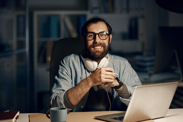 Portrait of happy hipster young bearded man in eyeglasses sitting at desk with laptop in night...