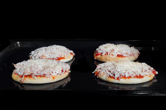 Four uncooked mini pizzas with ham, sause, cheese on tray in electric oven - front view, black background. Italian cuisine, homemade bakery, fast food, semi-finished products and raw concept
