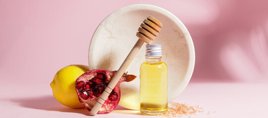 Bottle of natural cosmetic oil, honey dipper, lemon and pomegranate on pink background