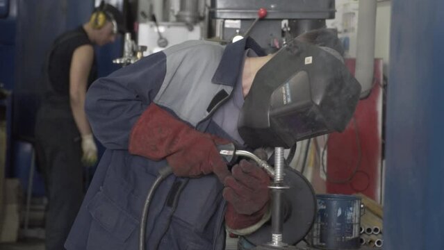 A factory worker in a protective mask welds metal. The man is welding. Welding with argon or electrode, using a welding machine. Sparks and flashes fly. The worker is facing the camera