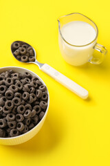Bowl with black cereal rings and jug of milk on yellow background