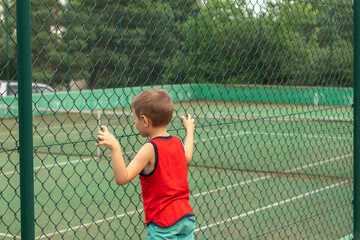 a little boy looks at the tennis court clinging his hands to a metal fence