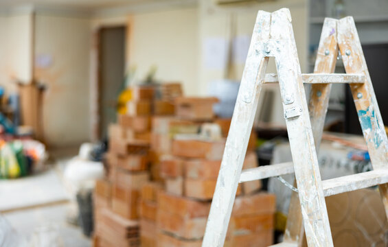 Ladder with stains of paint and plaster at indoor building construction site, various construction materials stacked on background