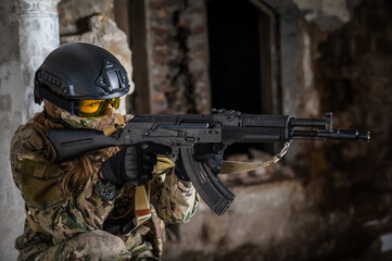 Fototapeta premium Portrait of a woman in a helmet and goggles with a machine gun in her hands. A female soldier in a camouflage uniform holds a weapon.