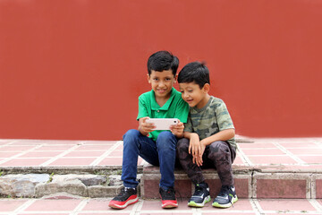 Two dark-haired Latino children who are brothers or friends who live in poverty play video games with a cell phone, make video calls and take selfies