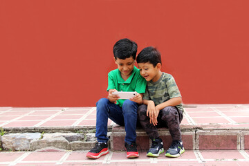 Two dark-haired Latino children who are brothers or friends who live in poverty play video games with a cell phone, make video calls and take selfies