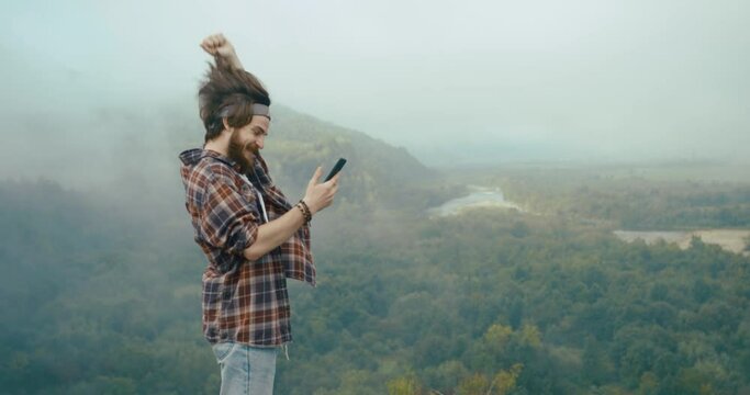 Young hypster man chats with someone on his phone standing on the top of the rock before beautiful mountain view covered with fog