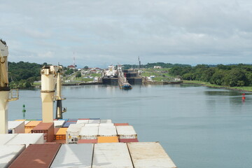 Container vessel with cranes approaching Gatun locks in Panama Canal form Atlantic Ocean to pass and reach Pacific Ocean. In the locks are other ships and tugs.