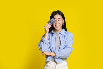 Happy excited young asian girl with credit, debit card. Asian girl in blue shirt holding mockup credit card isolated on yellow background. E-banking concept. E-commerce concept