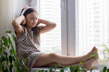 Cute little girl with wireless headphones sitting near window at home.