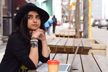 Portrait of young woman in fedora hat sitting at outdoor cafe with laptop computer daydreaming