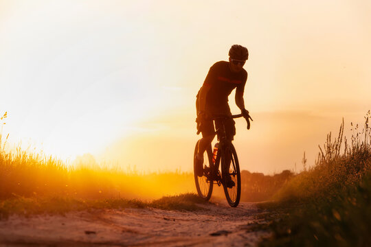 Silhouette young of cyclist on a gravel bike riding on a dust trail at sunset.
