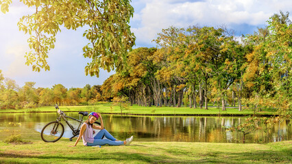 asian female biker sitting beside bicycle by lake in park relaxing after cycling for exercise for wellbeing life during autumn
