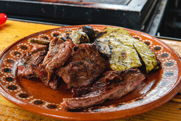 A delicious plate of meat and roasted nopales. Food on a clay plate. Wooden table and in the background a meat grill. Traditional dish of Mexico. Roasted fine cuts. Grilled top sirloin.