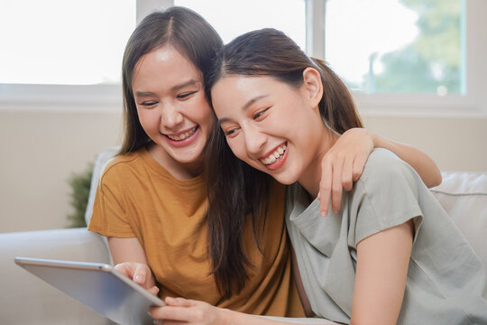 Happy lesbian, pleasure asian young two women, girl gay or close friend, couple love embrace, spending good time together, using tablet for entertainment on sofa at home. Activity of leisure, relax.