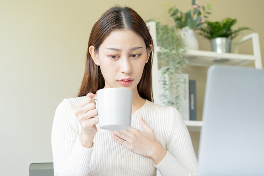 Palpitation, suffer asian young woman holding mug, drinking strong a cup of coffee, touching her chest after drink caffeine, tea while working from home, face expression stress, illness people.