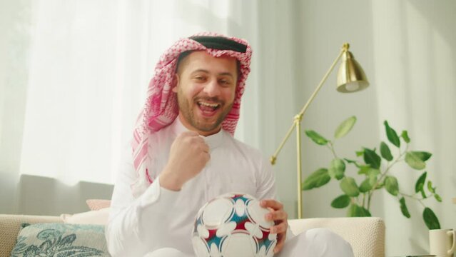Middle eastern football fan supporting favorite team, watching match on TV. Young man holding soccer ball. Wearing traditional Islamic male clothes. Worship and culture concept.