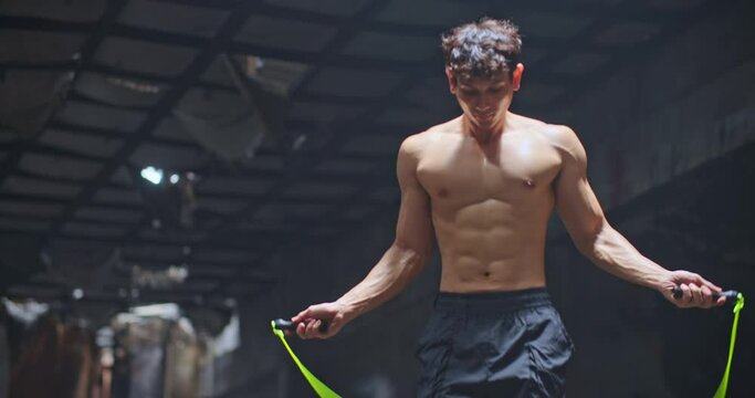 Young handsome Caucasian man boxer jump rope exercises in abandoned building hall gym under smoke light. Skipping man working out in cross training space in slow motion. Silhouette on dark background