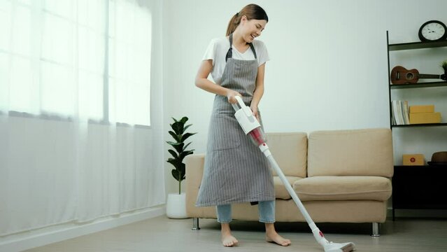 Beautiful housewife wearing an apron  plays music and dances while using a vacuum cleaner to clean the living room at house. Young woman is happy to clean home. Maid cleaning service.