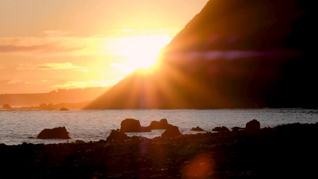 Beautiful, calm golden sunset with camera lens flare and glare, sun dipping behind mountain over ocean in New Zealand, Aotearoa