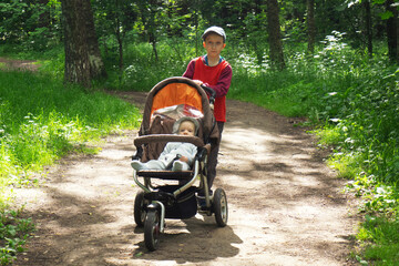 Young boy in red shirt and cap walking in forest with his toddler brother in pram. Childhood concept