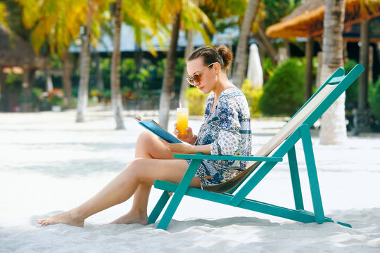 Happy woman sitting in deck chair reading book at tropical beach  in resort.