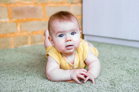 Portrait of smiling little Caucasian baby girl in yellow dress with wooden teething toy. Child trying to crawl on a floor.