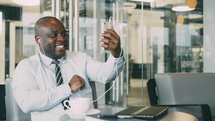 Naklejka na ściany i meble Portrait of African American businessman in formal clothes smiling and talking to his family through smartphone webcam in a modern cafe. He waving his hand happily having earbuds in his ears