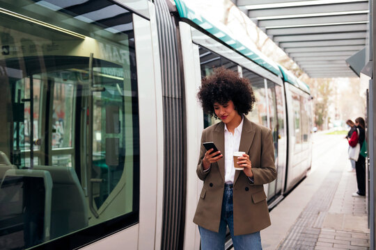 Young Business Woman Checking Her Phone While Waiting For The Tram To Arrive, Concept Of Technology And Urban Lifestyle