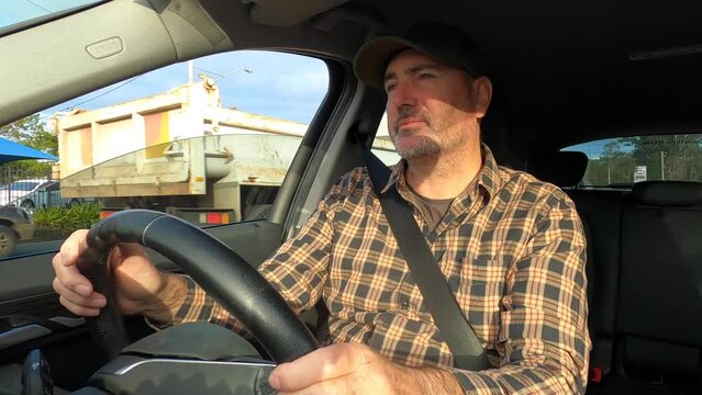 A man wearing a cap squints and uses a sun visor while driving in a country town towards the sun.