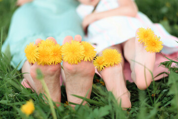 Close-up of women's and children's bare feet with dandelion flowers between the toes.Mom and daughter sitting on the grass on a summer day.Summer,closeness to nature,simple slow living.Selective focus