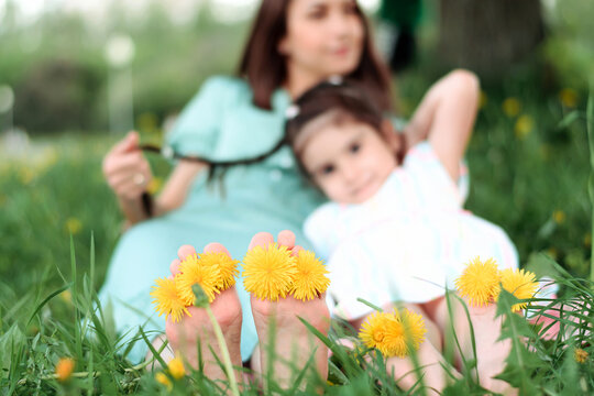 Blurry mother and daughter sitting on the grass in the park on a summer day.In the foreground are bare feet with dandelion flowers between the toes.Summer,closeness to nature,simple slow living.