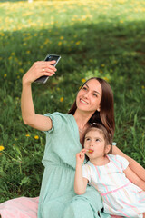 Obraz premium Caucasian mother and daughter kid use a smartphone in the park on a summer day.Mom and child talk on video call,make self-portrait picture together.Summer, technology,social media,family concept.