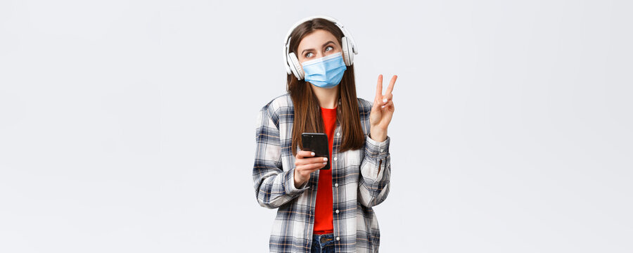 Social Distancing, Leisure And Lifestyle On Covid-19 Outbreak, Coronavirus Concept. Happy Cheerful Silly Girl In Medical Mask And Headphones, Show Peace Sign Smiling, Holding Mobile Phone