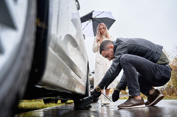 Charming blonde woman standing by automobile and holding umbrella while man placing car jack under...