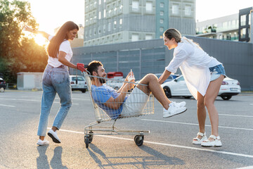 Three diverse friends having fun and riding shopping cart on parking lot near supermarket