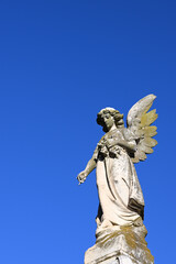 Worn and slightly mossy stone sculpture of an angel mournfully dropping flowers, with clear blue sky in the background