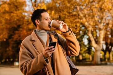 Young man using cellphone and drinking coffee in autumn park