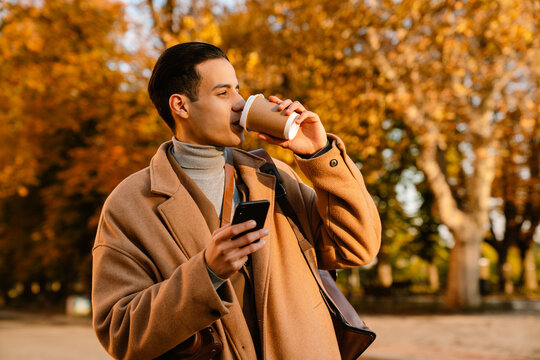 Young man using cellphone and drinking coffee in autumn park