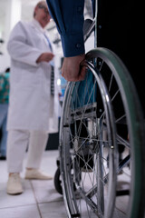 Fototapeta premium Closeup of older man hand using wheelchair in front of medical doctor for appointment in private hospital reception. Selective focus on senior man living with disability at medical clinic.