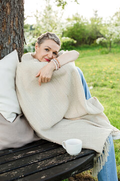 Woman wrapped in blanket sitting with coffee cup on bench by tree trunk
