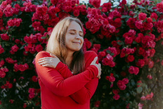 Smiling woman hugging self in front of rose plant