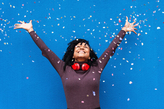 Happy woman wearing wireless headphones throwing confetti in front of blue wall