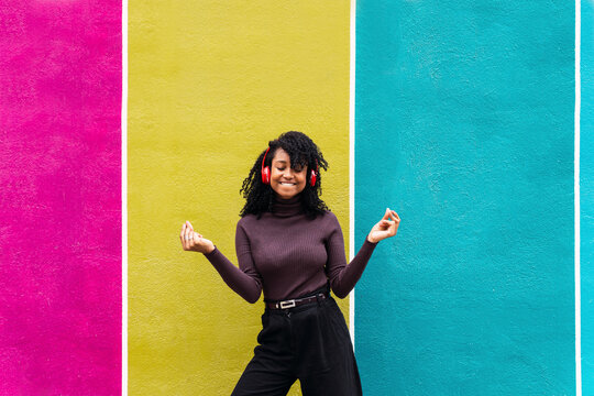 Smiling woman listening music through wireless headphones enjoying dance in front of multi colored wall