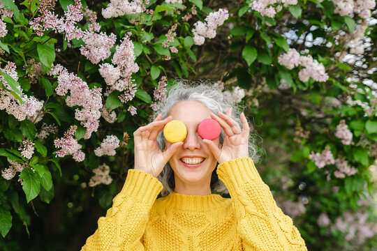 Happy woman covering face with macaroons in front of lilac at park