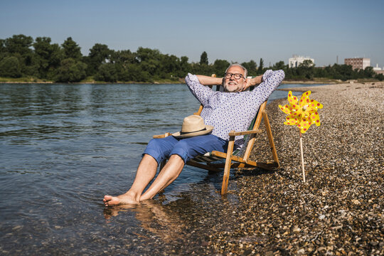 Smiling man with hands behind head sitting by paper pinwheel toy at riverbank