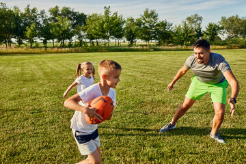 Father playing rugby with son and daughter at sports field on sunny day