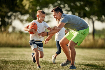 Cheerful man chasing son running with rugby ball at sports field