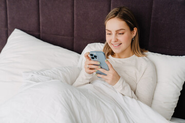 Young white blond woman smiling and using cellphone in bed at home